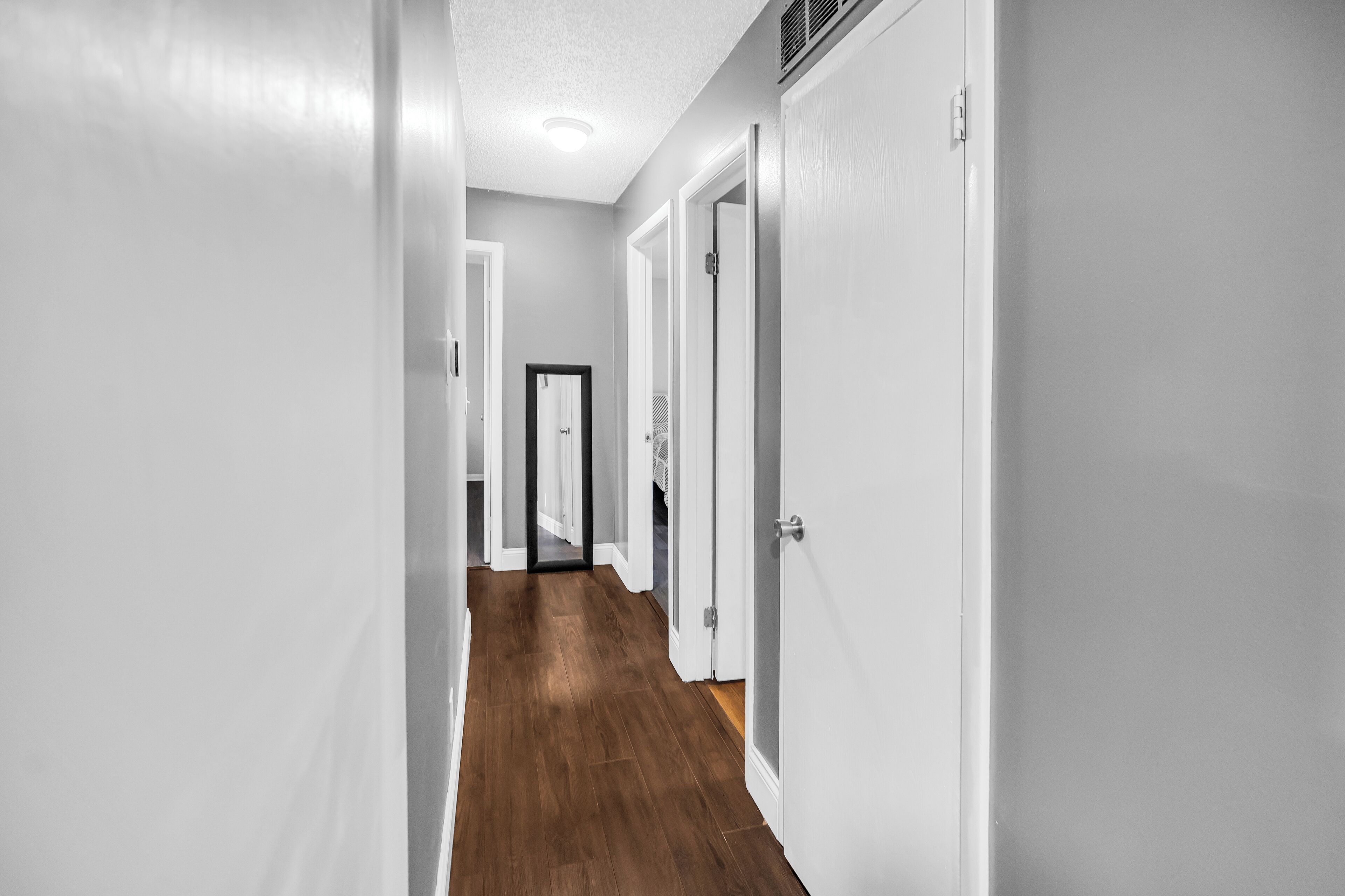 A hallway with white walls and wooden flooring, leading to a door with a mirror and a vent.