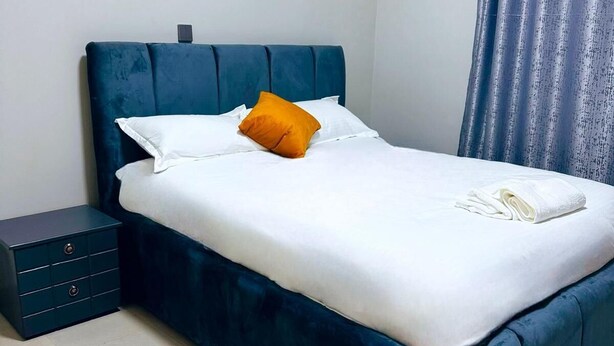 A neatly made bed with white sheets and a single orange pillow, a dark blue headboard, and a small bedside table with drawers. - 1 Bed-uganda-2people-wifi-closetocity-parking (Kampala)