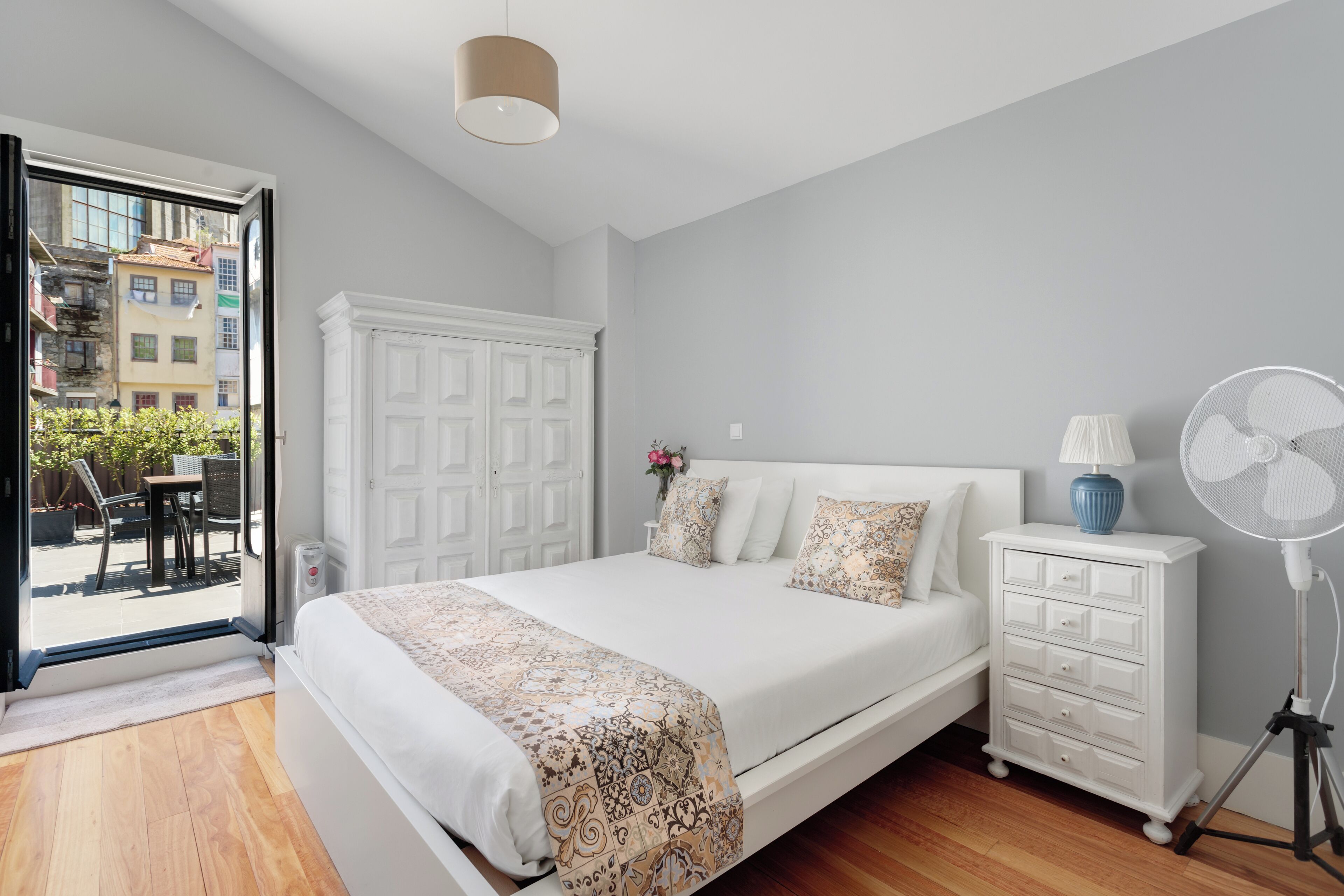 Un dormitorio con una cama grande, un aparador blanco, un ventilador de pie y vistas al exterior.