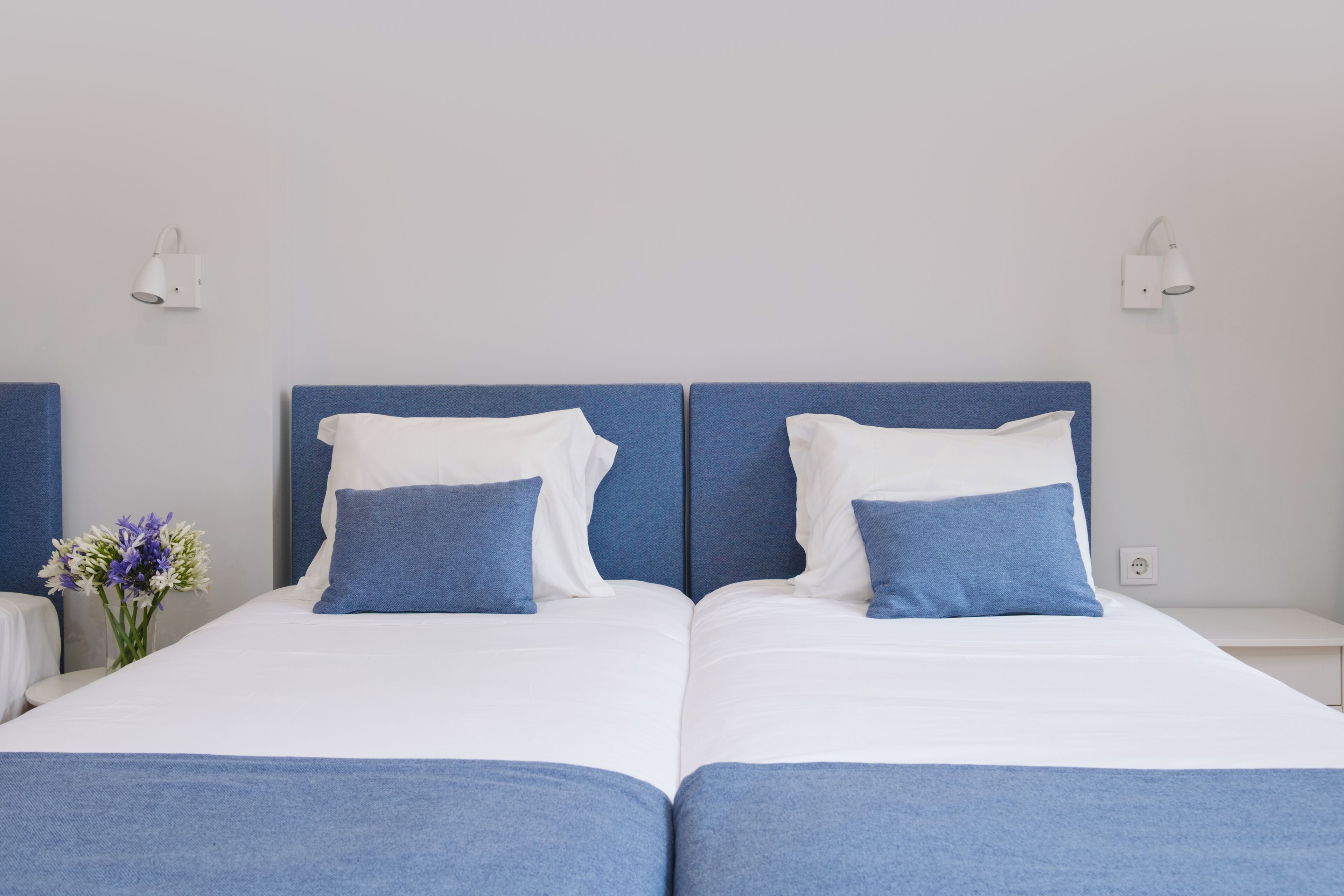 Uma cama de casal com roupa de cama branca e azul, duas mesas de cabeceira e uma luminária fixada na parede.