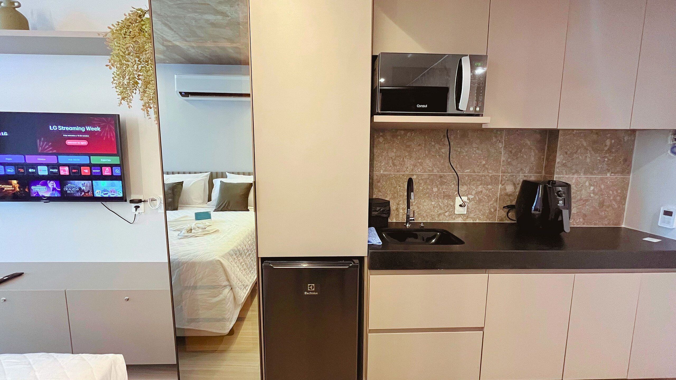 A compact hotel room with a bed, a small kitchenette, a TV displaying LG Streaming Week, and a bathroom with a sink and mirror.