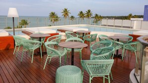 A rooftop pool area with green wicker chairs and tables, overlooking the sea.