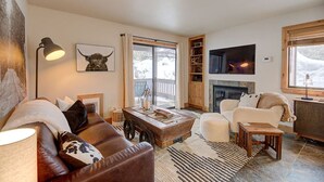 A cozy living room with a fireplace, leather sofa, and a large window with a view of the outdoors. - Pinetree Retreat - Hot Tub, Heated Pool & Sauna - 6 Min Walk to Dining & Lift (Park City)