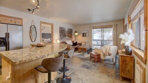 A modern kitchen with a granite countertop, bar stools, and a dining area with a sofa and coffee table. - Pinetree Retreat - Hot Tub, Heated Pool & Sauna - 6 Min Walk to Dining & Lift (Park City)