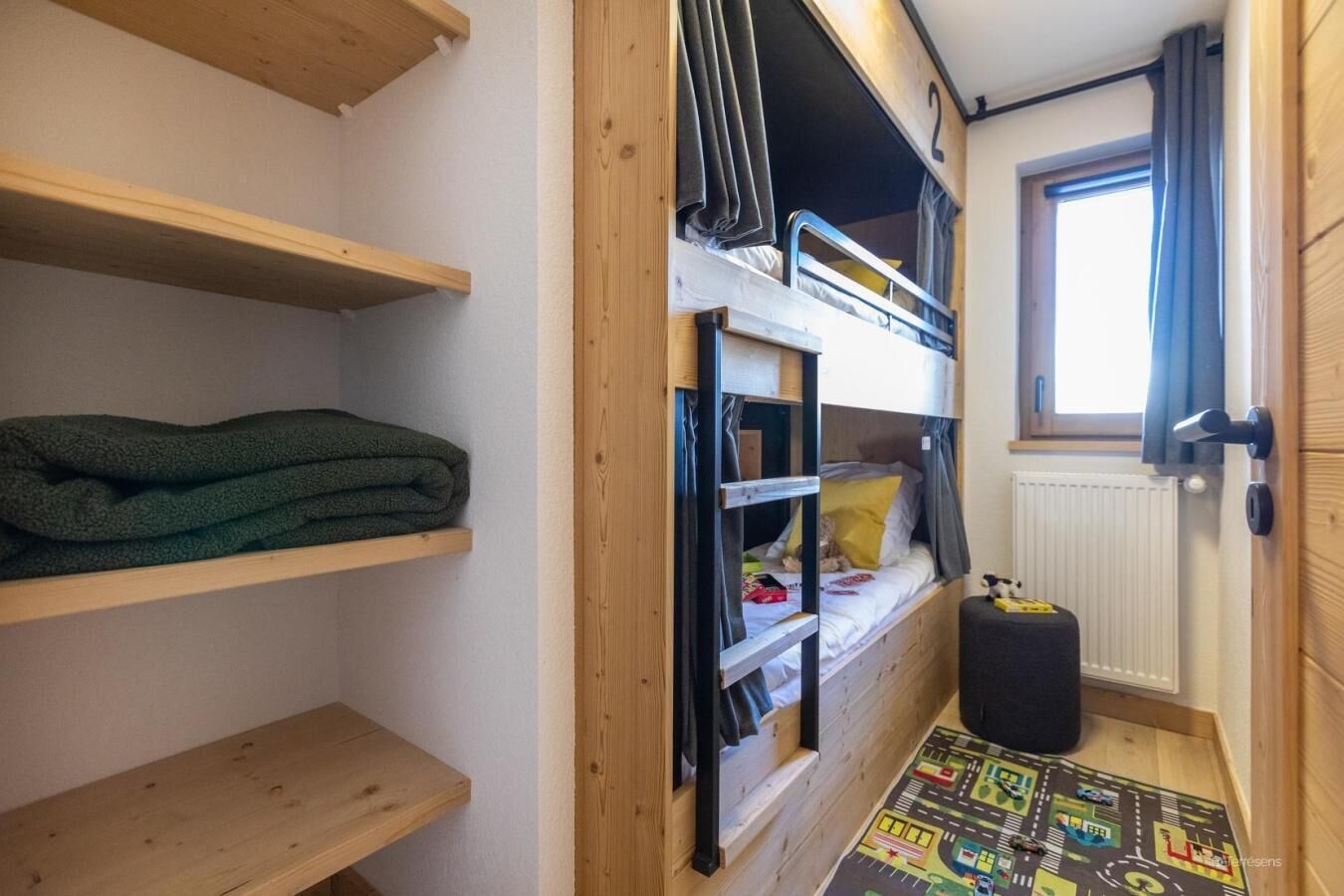 Une chambre de dortoir avec des lits superposés, des étagères en bois, un radiateur et une fenêtre avec des rideaux.
