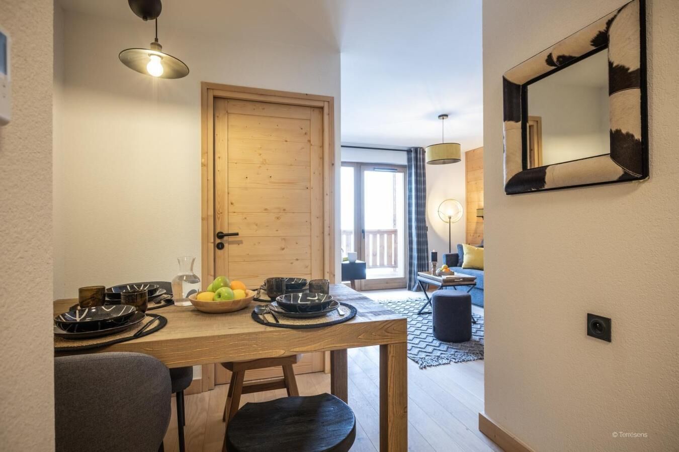 A dining area with a wooden table set for two, a bowl of fruit, and a mirror on the wall.
