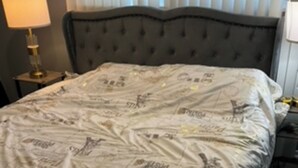A bed with a patterned bedspread, a bedside table with a lamp, and a window with blinds.
