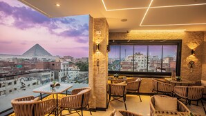A rooftop bar with wicker furniture, a large window with a cityscape view, and a wall-mounted light fixture. - Royal Pyramids View Inn (Giza)