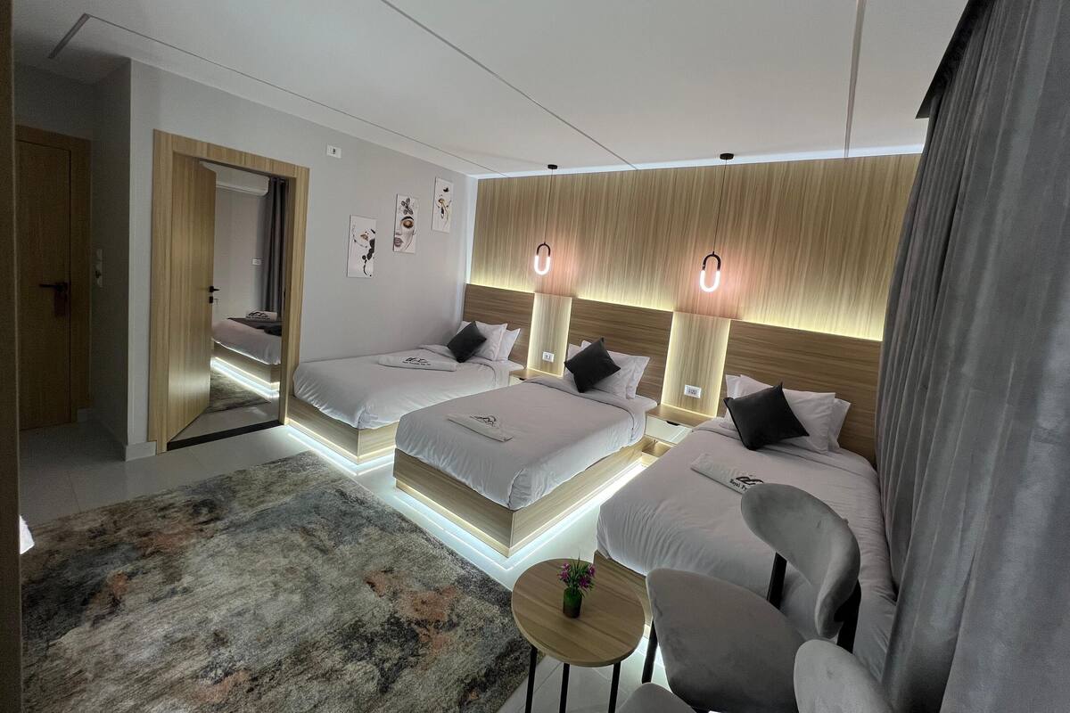 Ein modernes Hotelzimmer mit zwei Betten, einem Schlafsofa, einem kleinen runden Tisch und einem großen Spiegel.