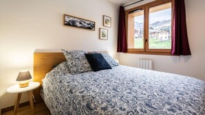 A bedroom with a bed, a nightstand, a lamp, a window with curtains, and two framed pictures on the wall. - 15m from ski slopes, 1st floor, view mountain, balcony, tv, ski locker, 49m², Valmorel (Valmorel)