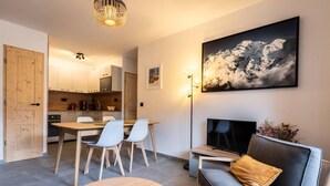 A modern living room with a sofa, a coffee table, and a TV. There is a dining area with a table and chairs. The room has a large window with a view of mountains. - 15m from ski slopes, 1st floor, view mountain, balcony, tv, ski locker, 49m², Valmorel (Valmorel)