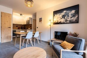 Ein modernes Wohnzimmer mit einer Couch, einem Couchtisch und einem Fernseher. Es gibt einen Essbereich mit Tisch und Stühlen. Das Zimmer verfügt über ein großes Fenster mit Bergblick.
