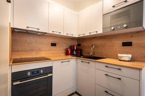 Eine kleine Küche mit weißen Schränken, einem schwarzen Backofen, einer Edelstahl-Dunstabzugshaube, einer Holz Arbeitsplatte, einem Spülbecken und einer Kaffeemaschine.