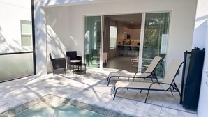 A modern outdoor patio with a swimming pool, a dining area with chairs, and a kitchen visible through glass doors.
