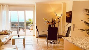 Un salon comprenant un canapé, une table basse et un coin repas avec des chaises et une table. Il y a également un balcon avec vue sur l’océan.