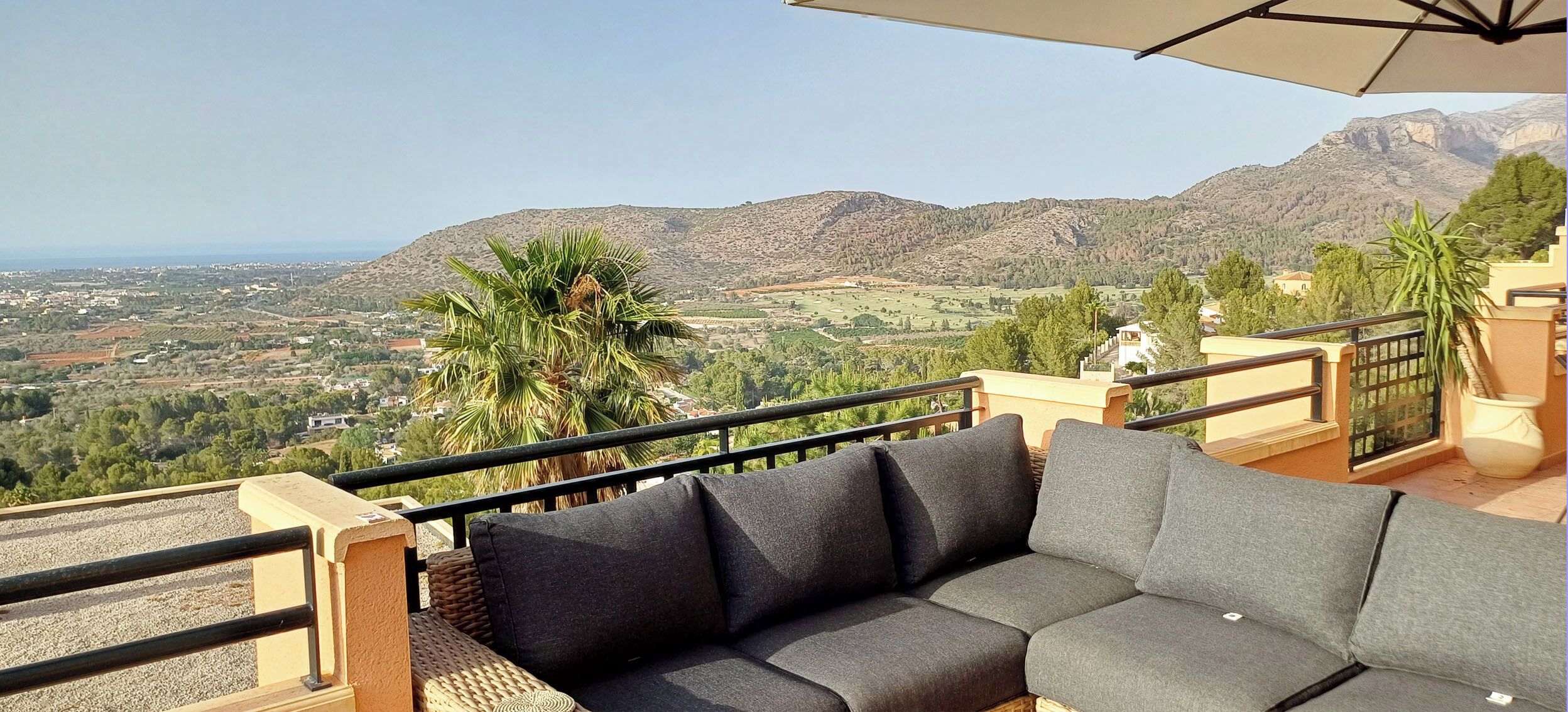 Ein Balkon mit einer Couch und einem Sonnenschirm, mit Blick auf eine gebirgige Landschaft und ein Dorf darunter.