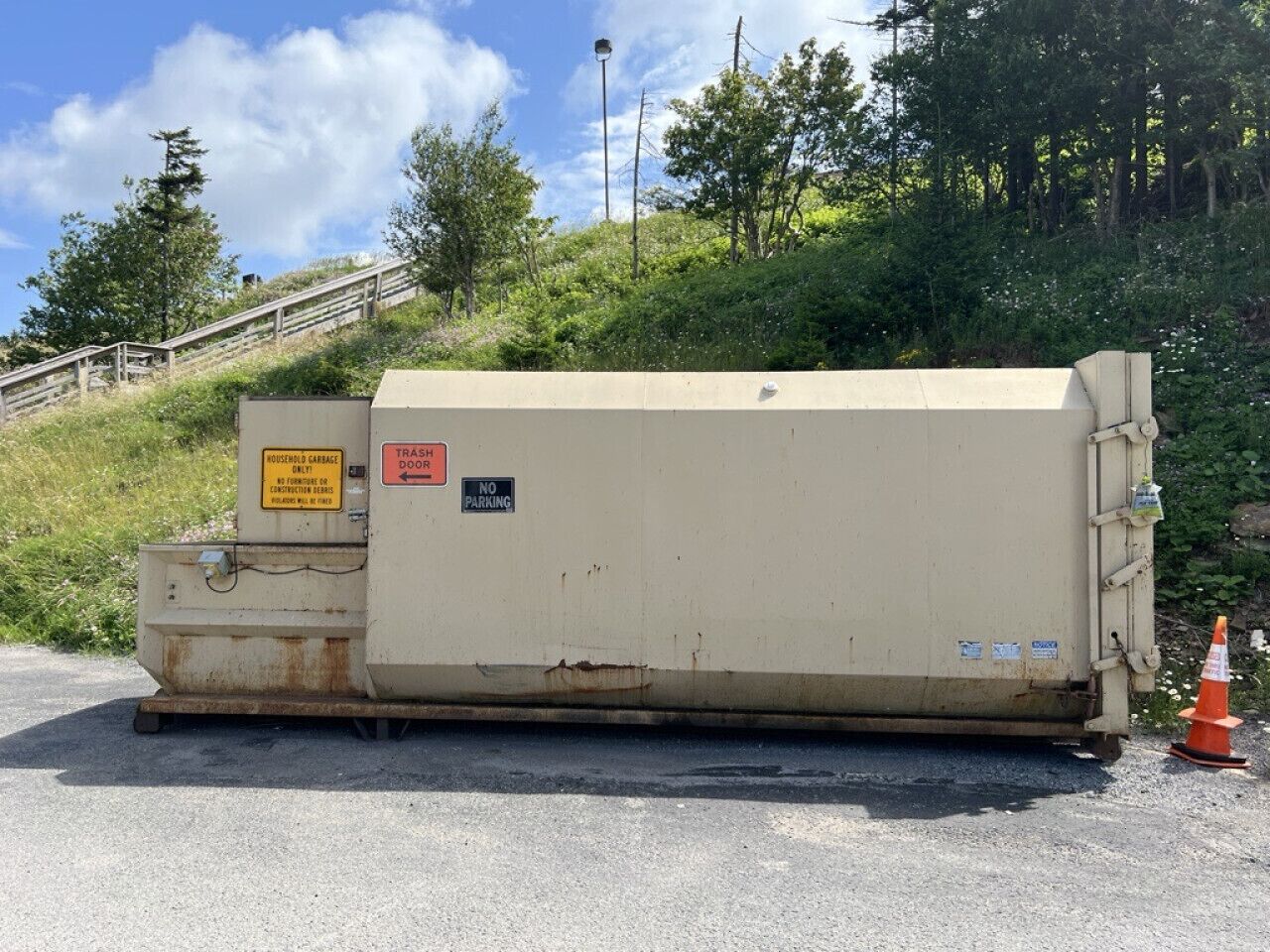 Une grande benne à ordures de couleur beige, avec une poubelle à proximité et un panneau « interdiction de stationner » sur une surface en béton. On aperçoit en arrière-plan une colline herbeuse et des arbres.