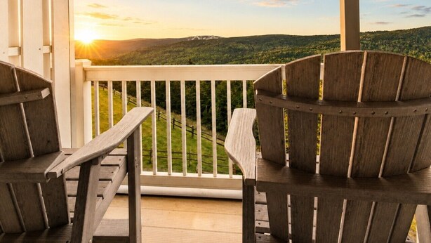 Une terrasse en bois avec deux fauteuils Adirondack, une balustrade blanche et une vue panoramique sur un paysage boisé au coucher du soleil.