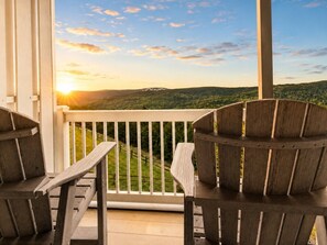 Une terrasse en bois avec deux fauteuils Adirondack, une balustrade blanche et une vue panoramique sur un paysage boisé au coucher du soleil.