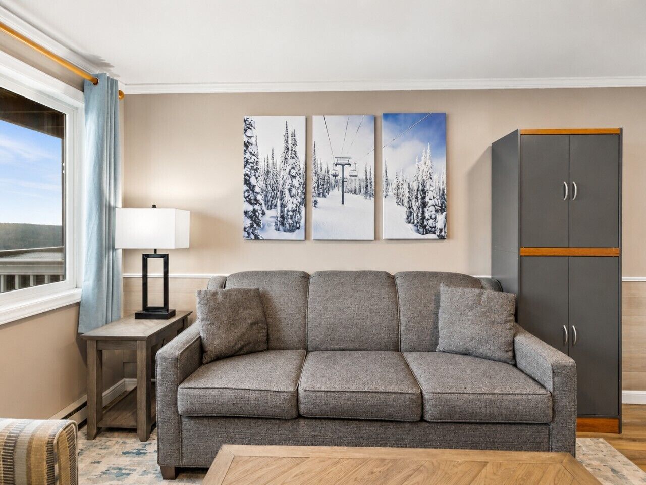 Een woonkamer met een grijze bank, een kleine tafel met een lamp, een kast, en drie ingelijste foto's aan de muur.