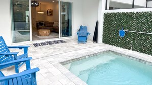 Un espace piscine extérieur moderne, avec une porte vitrée donnant sur un salon, une chaise bleue et une petite table.