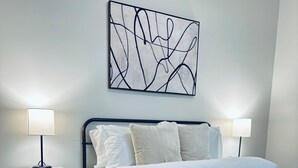 Quarto com cama, duas mesas de cabeceira, uma obra de arte abstrata na parede e chão de madeira.