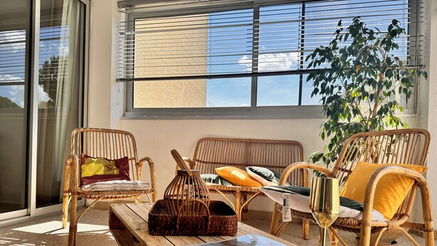 Una habitación iluminada por el sol con muebles de mimbre, incluyendo dos sillones y un banco, una cesta de mimbre, y una revista sobre el banco.