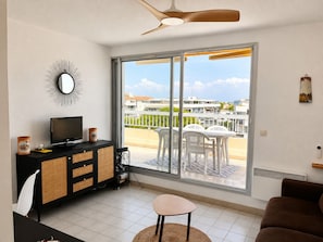 A modern living room with a flat-screen TV, a black entertainment unit, a dining area with white chairs, and a large sliding glass door leading to a balcony with a view of buildings and greenery.