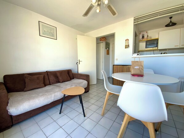 A living area with a brown sofa, a small round table, and a white chair. There is a door leading to another room, a ceiling fan, and a framed picture on the wall.