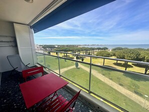 A balcony with a red table and chairs, overlooking a park and the sea.