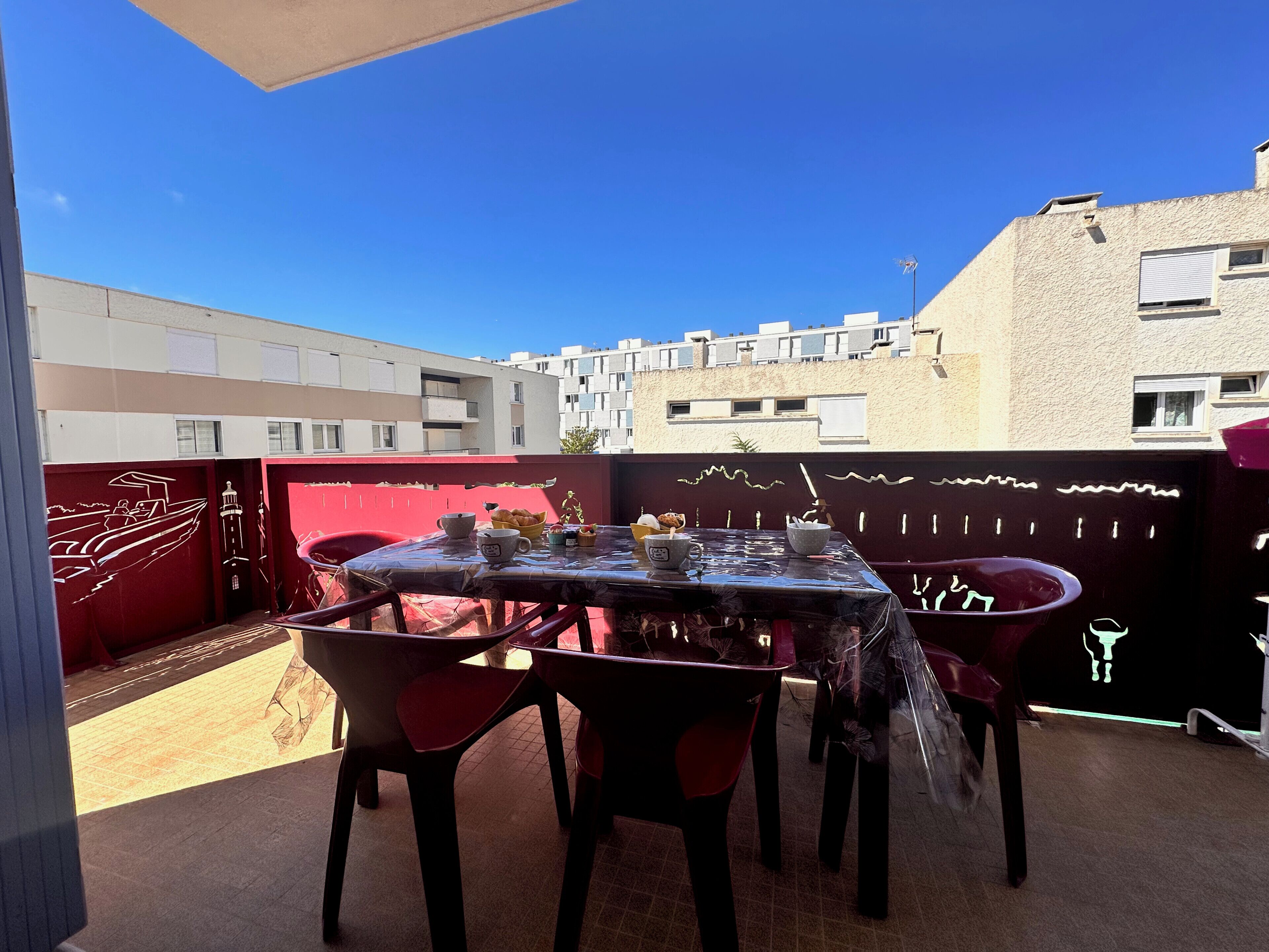 Un espace extérieur abrité, aménagé pour les repas, avec une table et des chaises, une chaise rouge et une table sur laquelle se trouvent divers objets.