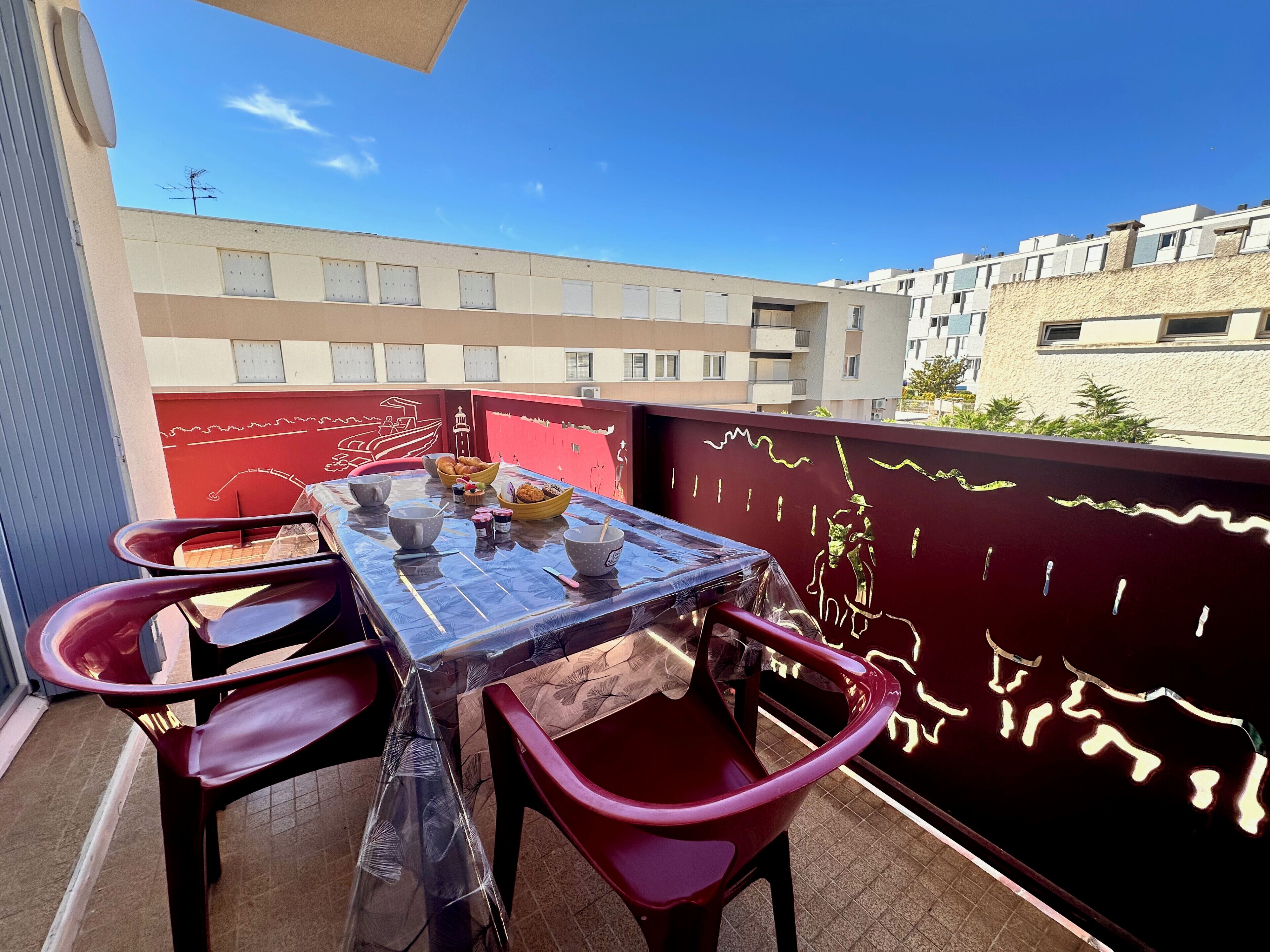 Ein Balkon mit Tisch für zwei Personen, bedeckt mit einer Schutzplane, und roten Stühlen.
