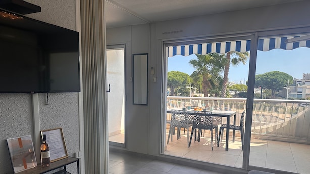 Un salon moderne avec un téléviseur, un certificat encadré et une petite table sur laquelle se trouve une bouteille. Il donne sur un balcon équipé d’une table et de chaises, d’où l’on peut admirer des palmiers et des immeubles.