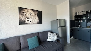 Un salon moderne avec un canapé gris, un grand tableau encadré représentant un lion et un coin cuisine doté d’un réfrigérateur et de meubles de rangement en acier inoxydable.