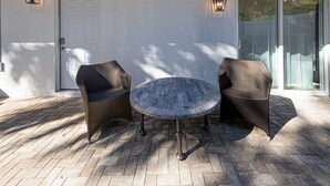 Patio esterno con due poltrone in vimini, un tavolo rotondo, una porta bianca e due lanterne fissate alla parete.