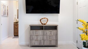 Un soggiorno moderno con una TV a schermo piatto montata a parete, un mobile contenitore in legno con cassetti e una pianta in vaso con fiori gialli.