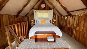 Ein gemütliches Schlafzimmer im Dachgeschoss mit schrägem Holzboden, einem Bett mit grau-weiß gemustertem Überwurf, einem Nachttisch aus Holz und einem Korb, der von der Decke hängt.