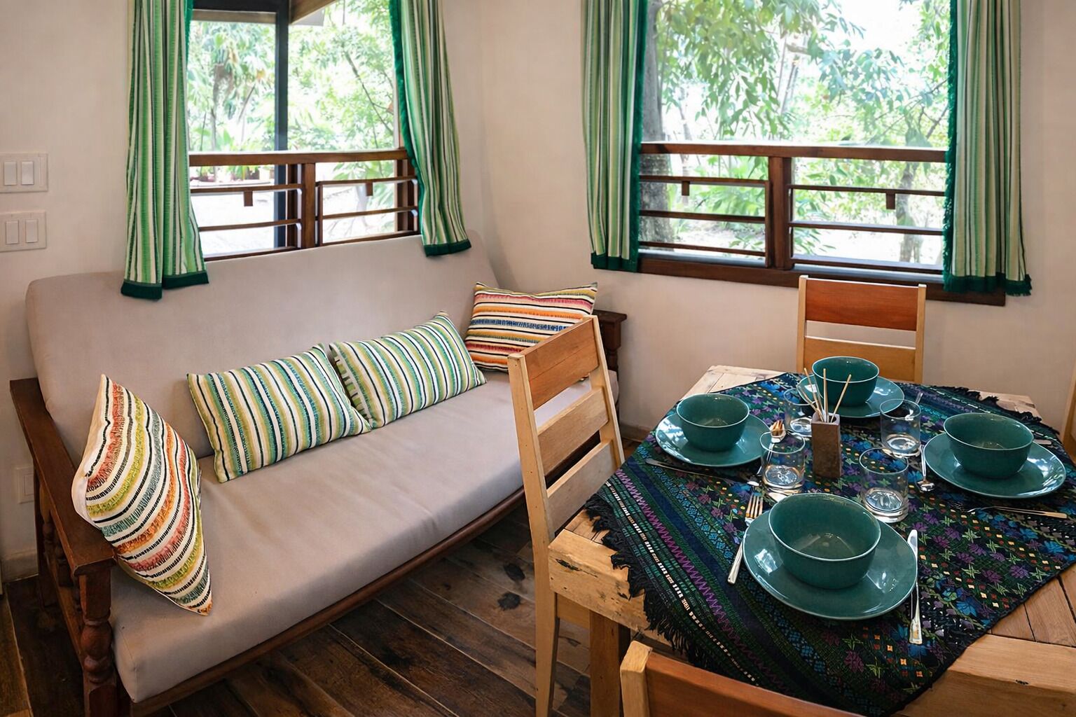 A room with a sofa, a dining table set with green plates and bowls, and a view of greenery outside.