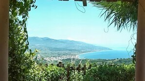 Vista a través de un arco de madera, con abundante vegetación, sobre un paisaje costero con montañas y un mar azul cristalino.