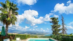 Una piscina rectangular con agua azul cristalina, rodeada por una terraza con baldosas. Hay sillas reclinables blancas y un paraguas verde a la izquierda. El fondo muestra un cielo despejado con algunas nubes y una vista distante de montañas y la costa.