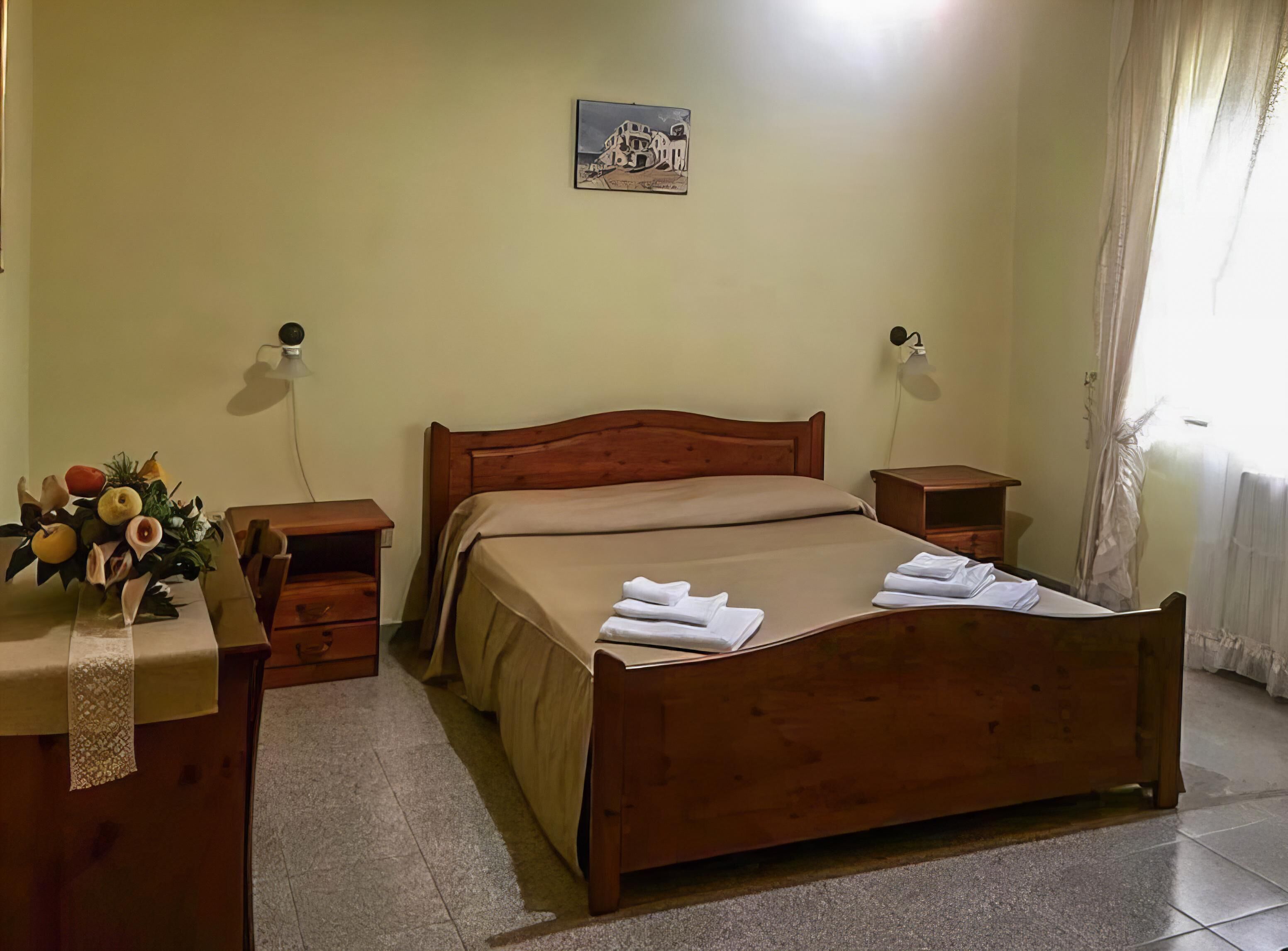 A neatly arranged bedroom with a wooden bed, a bedside table, a nightstand, a wall-mounted lamp, a framed picture, and a vase with flowers.