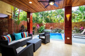 A covered outdoor area with wicker furniture, a pool, and lush tropical plants.