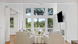 A spacious living room with a large window, a fireplace, and a flat-screen TV mounted on the wall. - Beautiful Bluebill Home on Kiawah, Private Pool, Pond and Golf Views! (Kiawah Island)