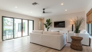 A spacious living room with a large white sofa, a TV mounted on the wall, and a sliding glass door leading to an outdoor area.