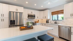 A modern kitchen with white cabinetry, stainless steel appliances, a central island with seating, and a window with blinds.