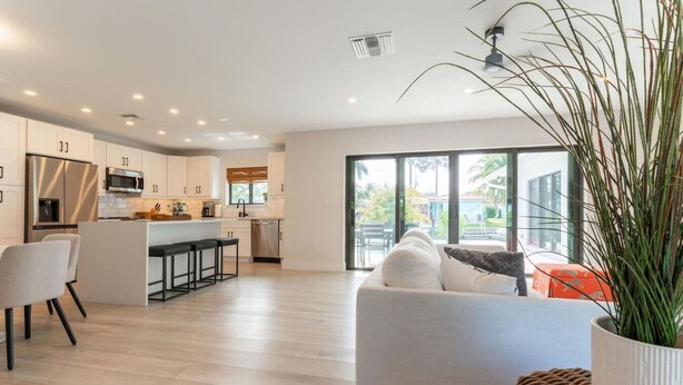 A modern kitchen with white cabinetry, a central island with bar stools, and a dining area with a white sofa and a plant.