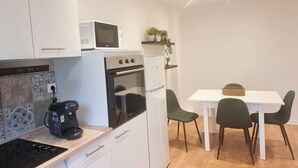 Cocina compacta con gabinetes blancos, microondas, cafetera y refrigerador. Hay una mesa de comedor con sillas y una pequeña planta en maceta sobre una repisa.