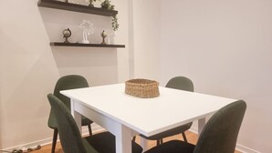Un comedor con una mesa blanca, cuatro sillas verdes y una cesta tejida sobre la mesa. Hay estantes encima de la mesa con plantas y adornos.
