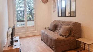 Una sala de estar acogedora con un sofá color beige, una mesita, un televisor y un ventanal con vistas a árboles y una cerca.