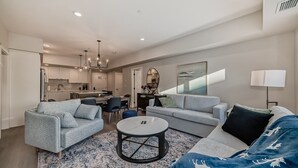 A modern living room with a sofa, armchair, and coffee table. There is a dining area with chairs and a table. The kitchen is visible in the background.